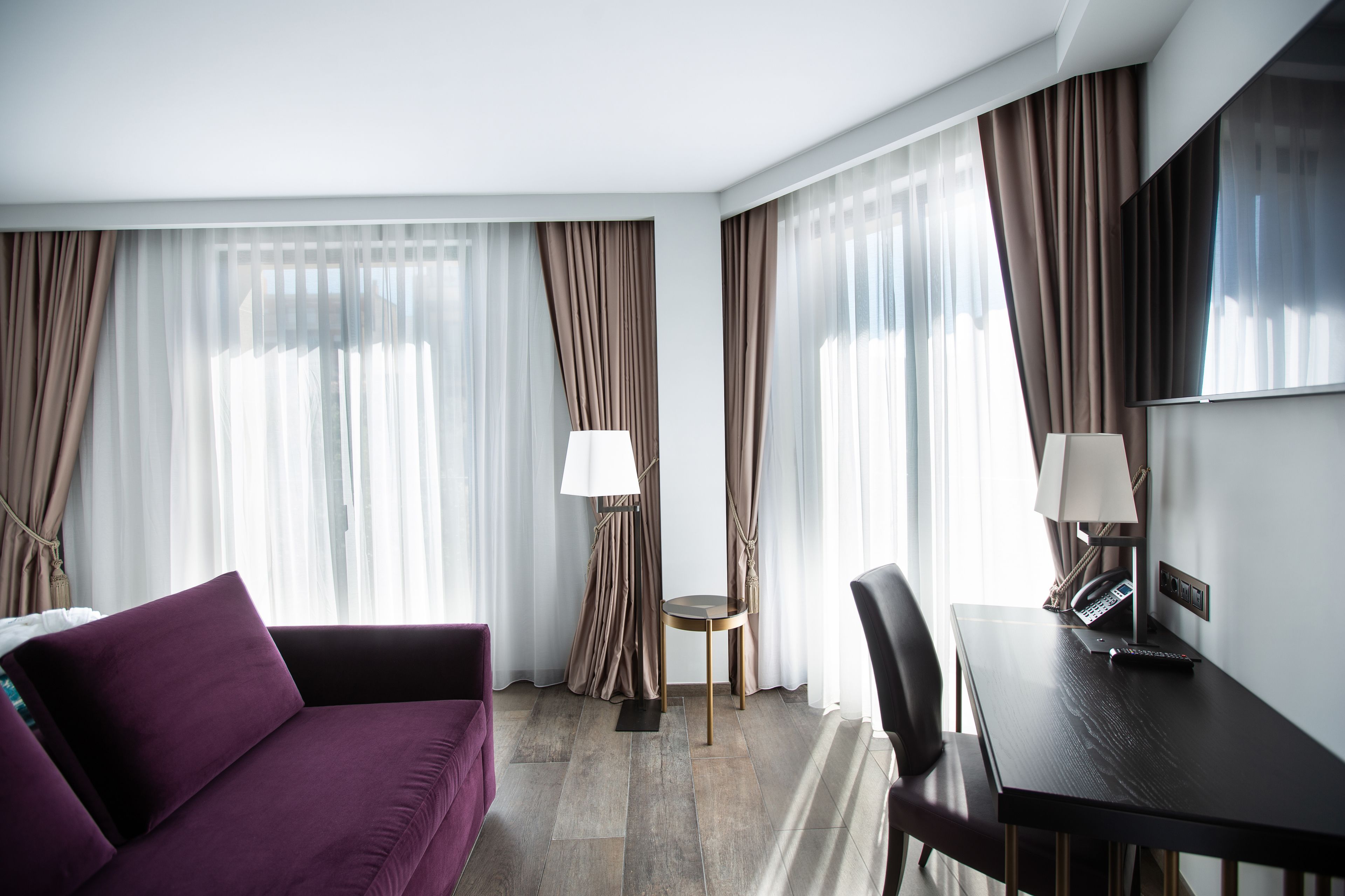 Quarto de hotel com sofá roxo, escrivaninha com abajur e uma cadeira.