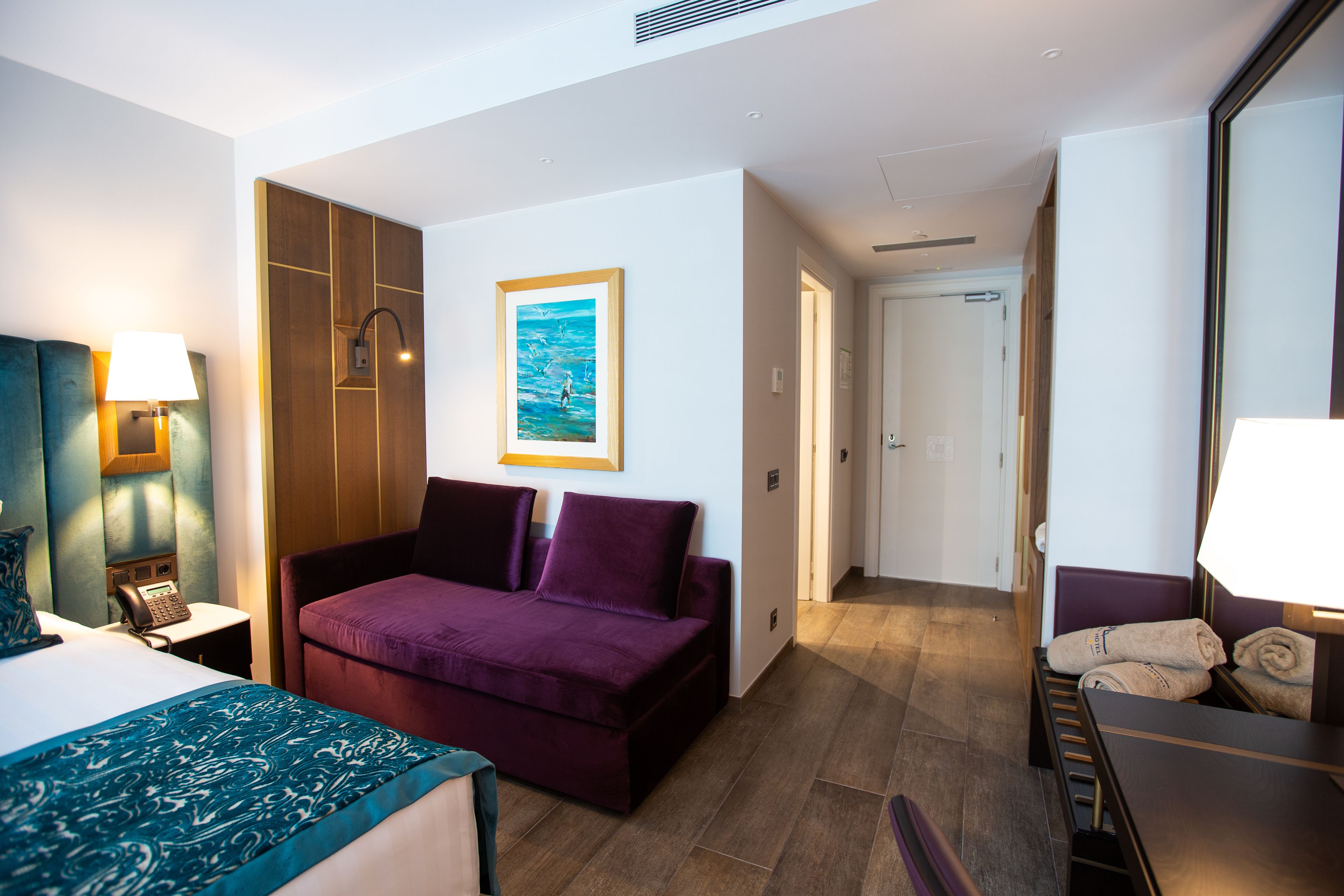 Een hotelkamer met een bed, een paarse bank, een houten kledingkast, een schilderij aan de muur, en een badkamer zichtbaar door een geopende deur.