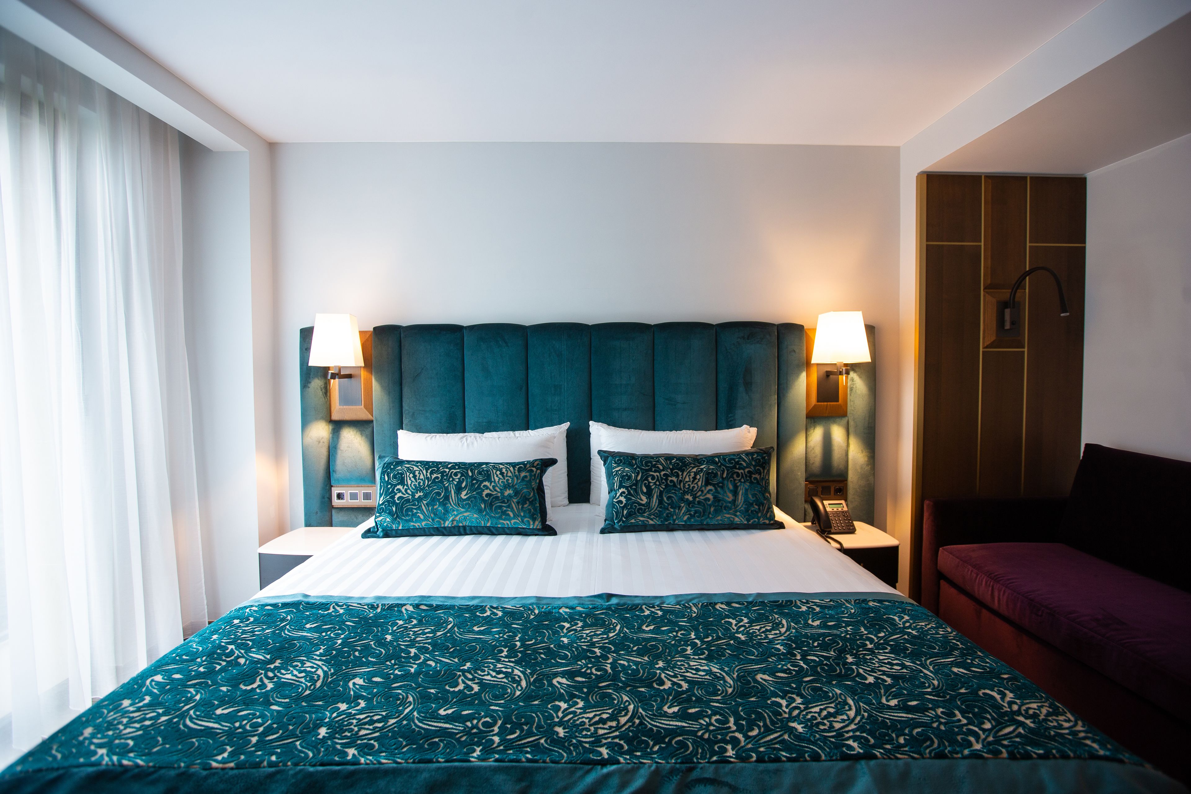 Habitació d'hotel amb un llit gran, dues làmpades laterals, un armari de fusta i una finestra amb cortines.