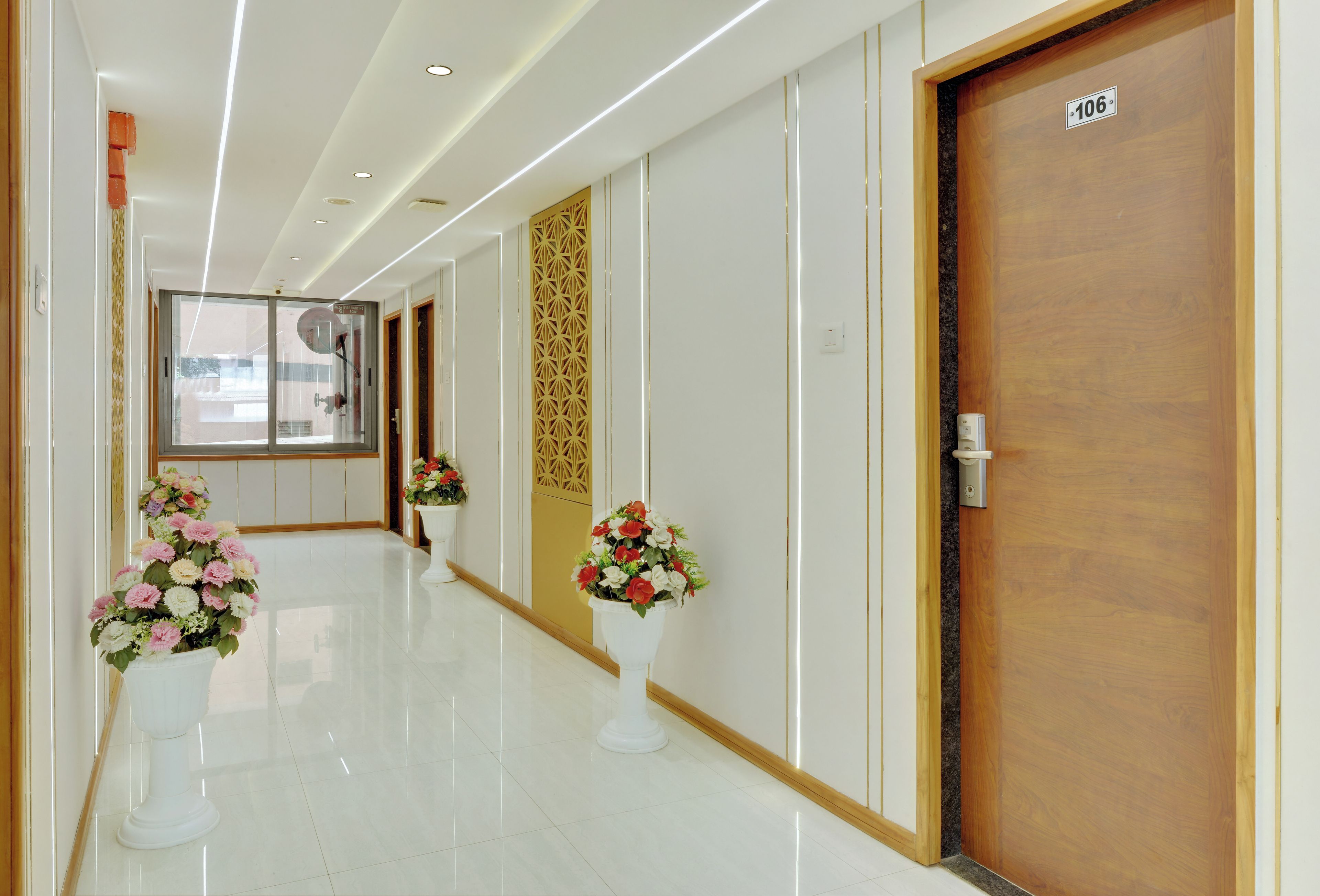 A hotel corridor with a wooden door labeled '106', a glass door with a view, and potted plants along the walkway.