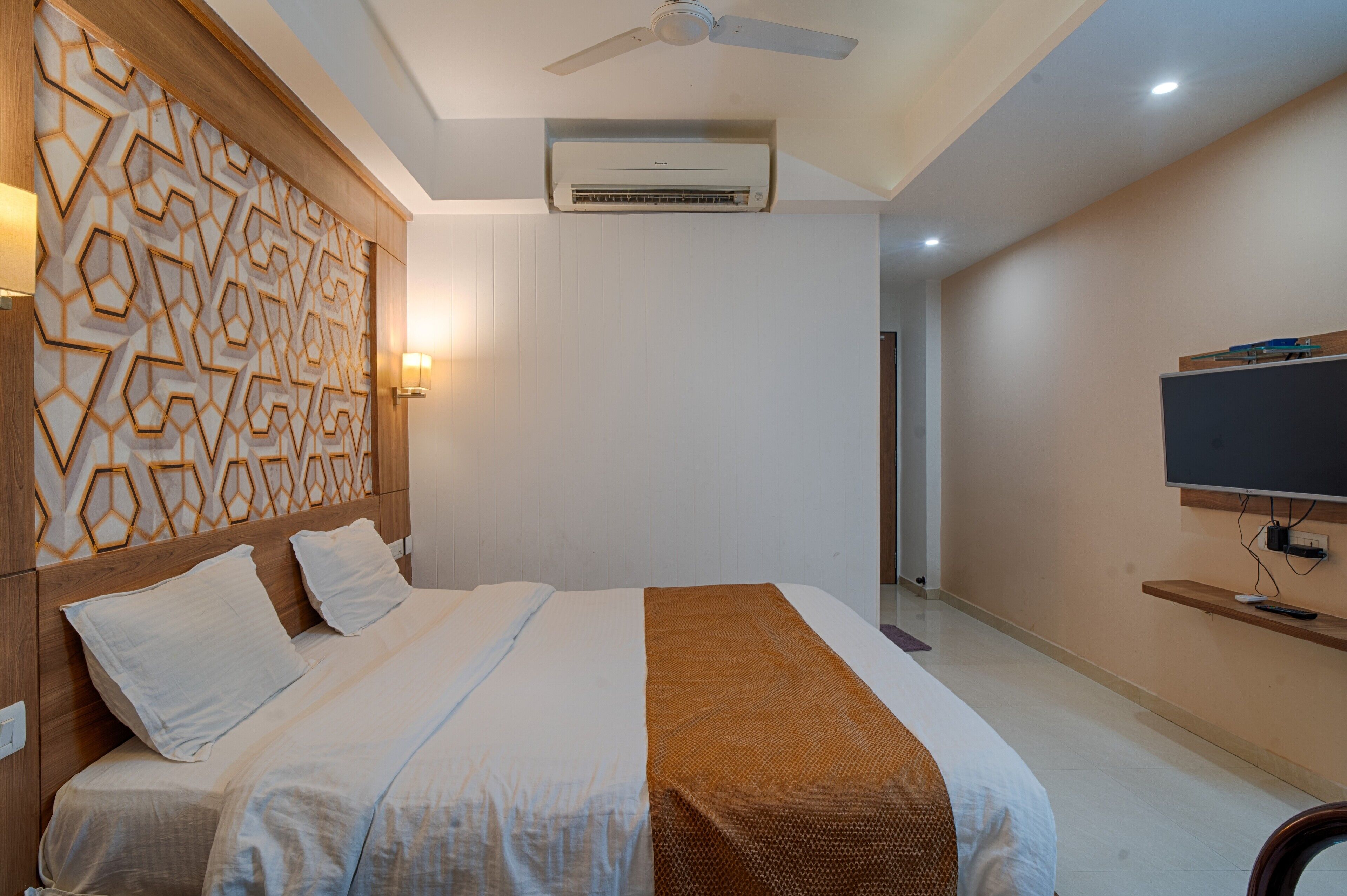 A modern hotel room with a large bed, a ceiling fan, an air conditioning unit, a TV on a stand, and a decorative wall panel.