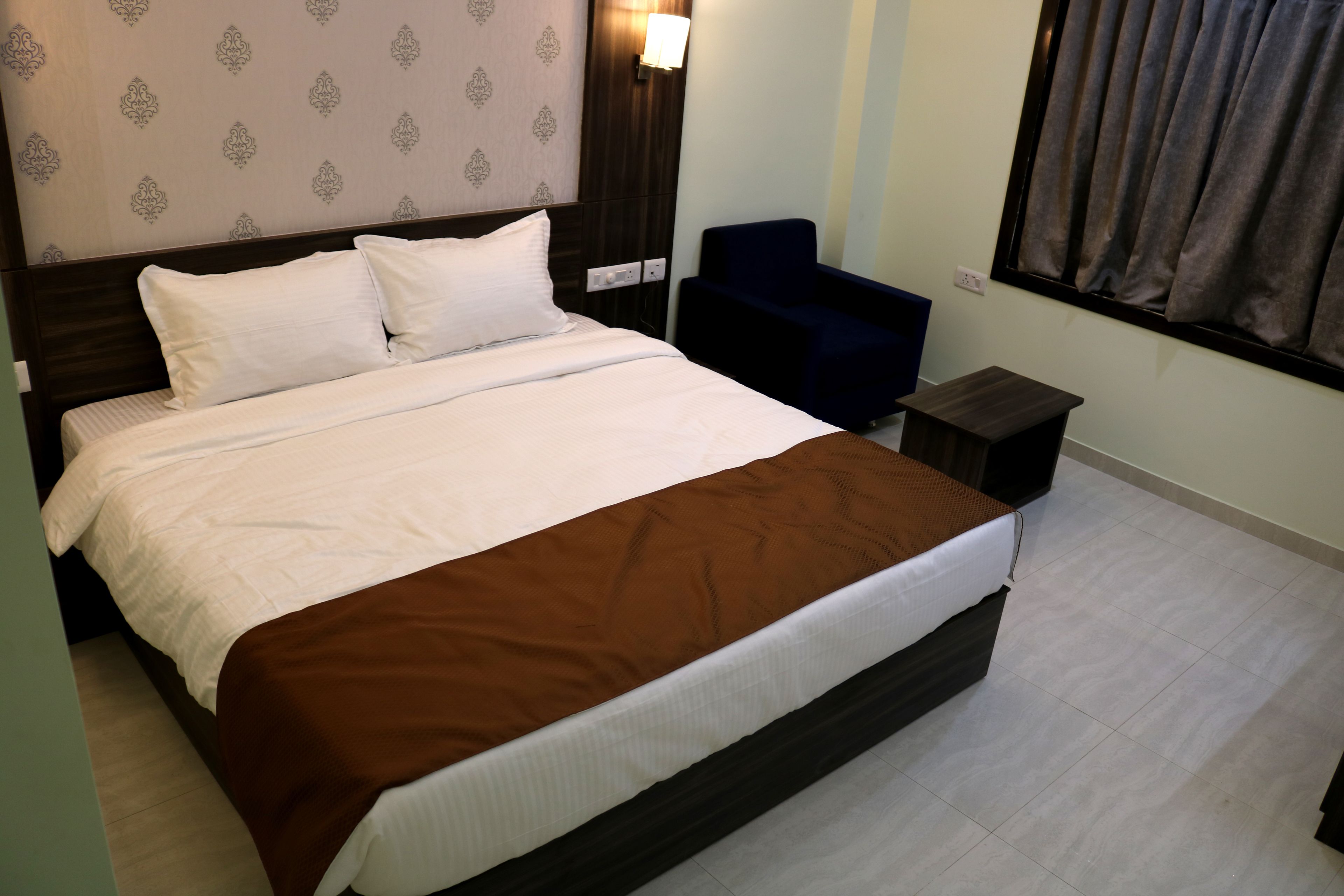 A neatly made bed with white and brown bedding, a dark headboard, a bedside table, a chair, and a window with curtains.