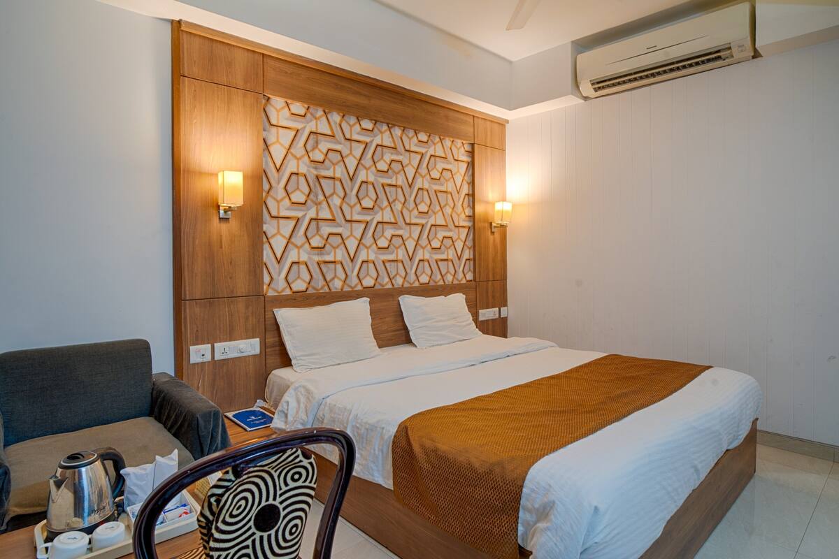 Une chambre d’hôtel avec une tête de lit en bois, deux lampes de chevet, un lit avec du linge de maison blanc, un plaid marron, un canapé gris, une table basse avec une bouilloire et une chaise à motifs.