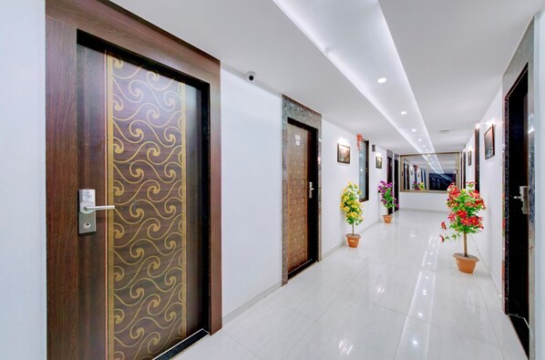 Corredor de hotel com porta de madeira com padrão decorativo, plantas em vasos e piso ladrilhado.