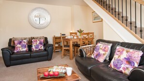 A living room with a black leather sofa, a wooden coffee table with apples and a white cup, a dining table with chairs, and a staircase with a wooden railing.
