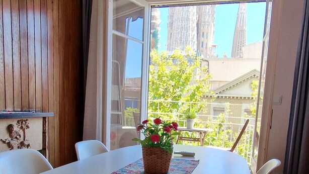 A dining area with a white table, a woven basket with red flowers, and a view of buildings and trees through a large window.