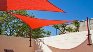 Un espacio al aire libre iluminado por el sol con dos sillones de descanso, una mesita con una jarra de vidrio y vasos, y un lavamanos blanco adosado a una pared.