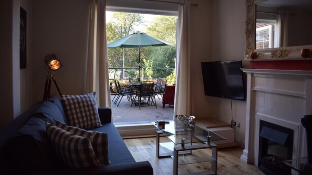 A living room with a sofa, TV, fireplace, and a view of an outdoor patio with a table and chairs. - Magma Flat (London)