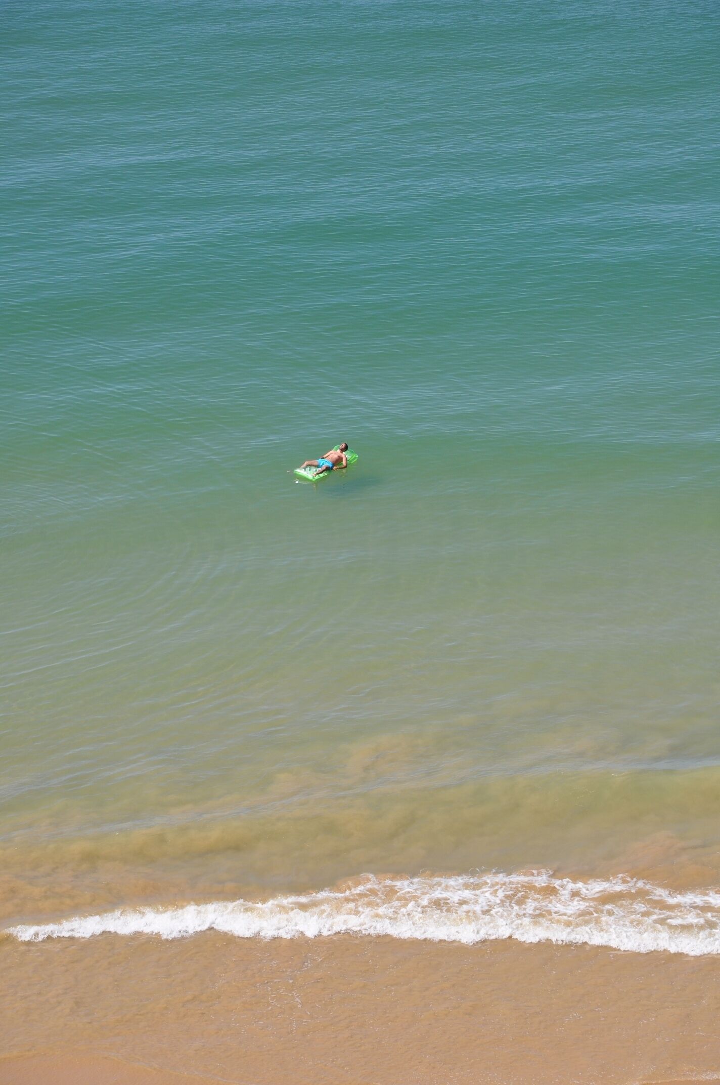 A person lying on a green float in calm, clear water near a sandy beach.