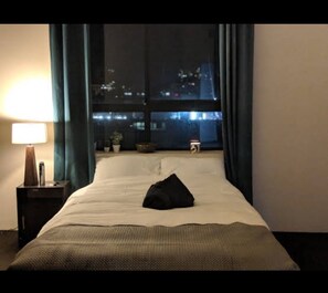 Una habitación de hotel con una cama, una mesita de noche, una lámpara, una ventana con cortinas y vistas a la ciudad de noche.