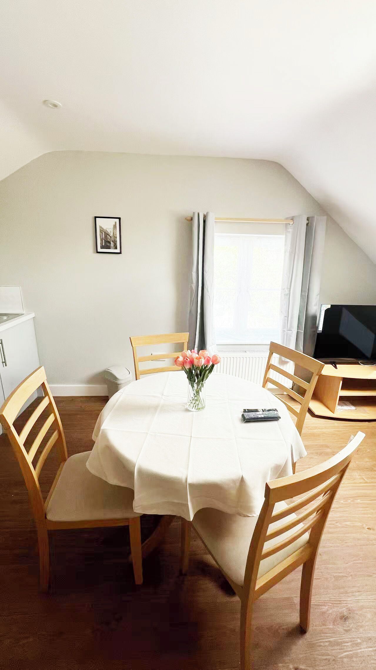 A dining area with a round table, chairs, a vase of flowers, a television, and a window with curtains.