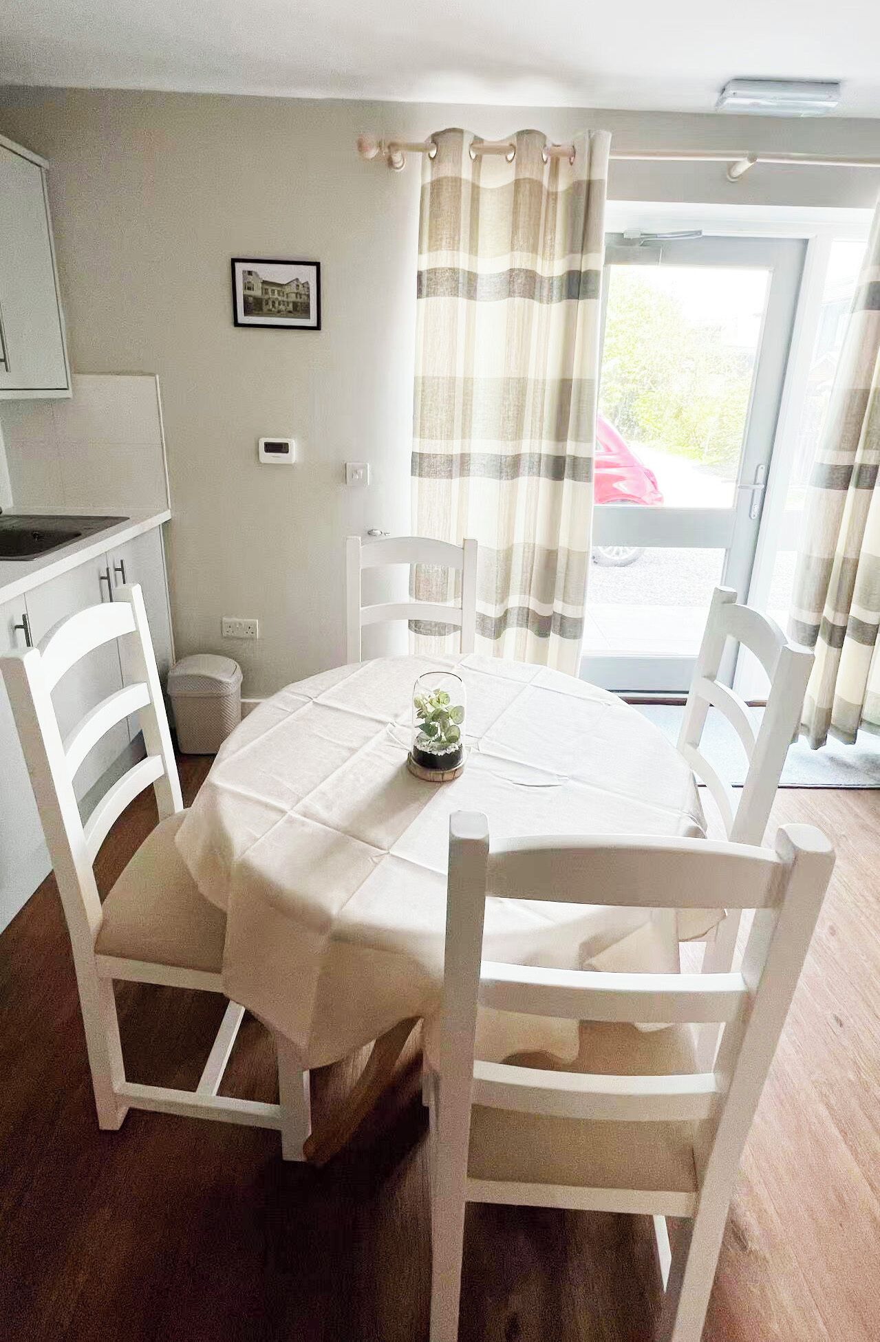 Un coin repas avec une table dressée pour quatre personnes, des chaises blanches, une petite plante en guise de décoration centrale et une vue à travers une porte vitrée.