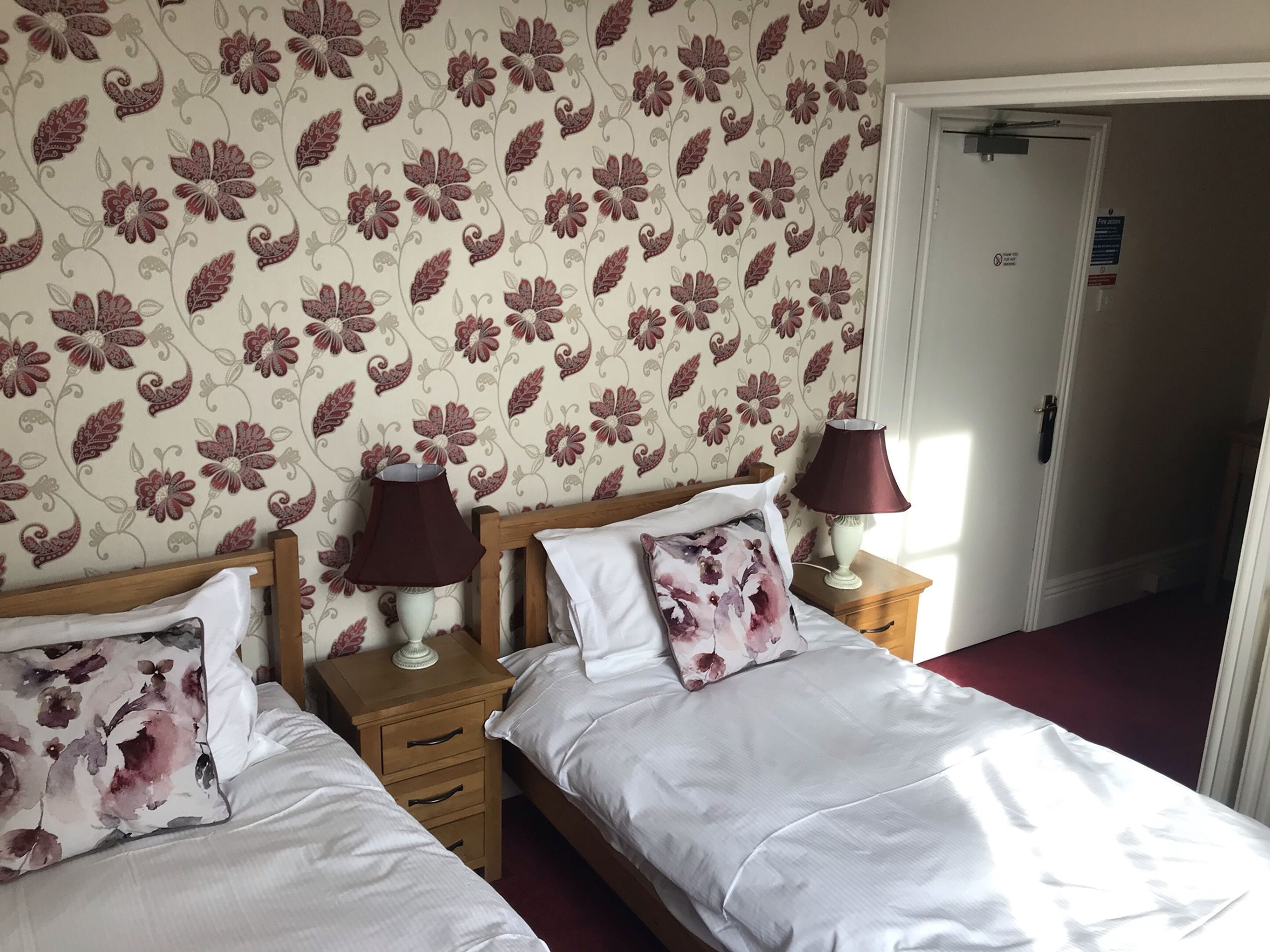 A hotel room with two beds, floral wallpaper, bedside lamps, and a door with a sign.