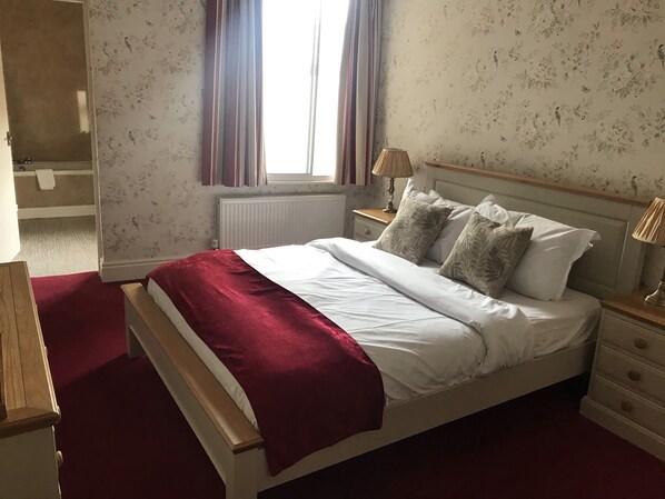 A bedroom with a bed, a nightstand, a lamp, a window with curtains, and a radiator.