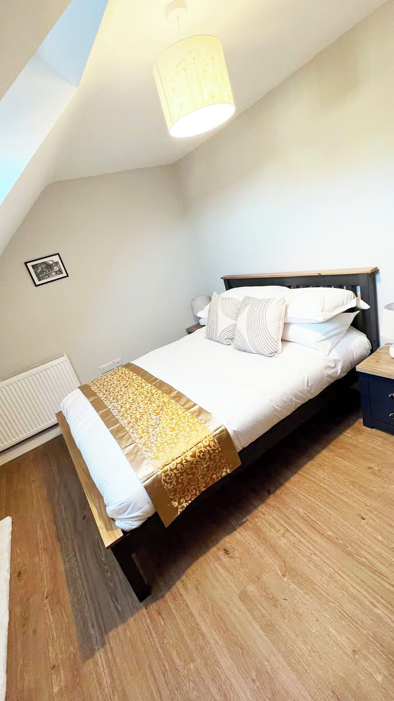 A bedroom with a bed, a bedside table, a lamp, a picture on the wall, and a ceiling light.