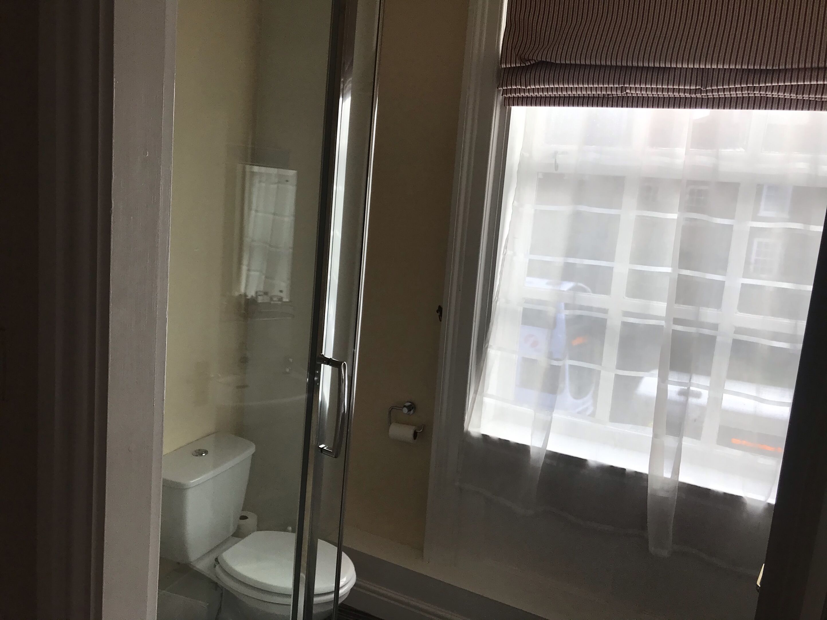 Une salle de bain avec une douche à parois de verre, des toilettes et une fenêtre ornée d’un rideau à motifs géométriques.