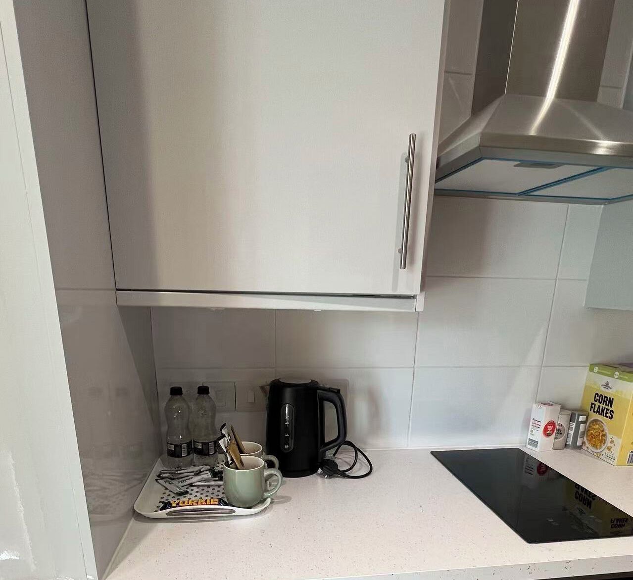 A kitchen with a stainless steel range hood, a kettle, a mug, a tray with utensils, and a box of corn flakes.