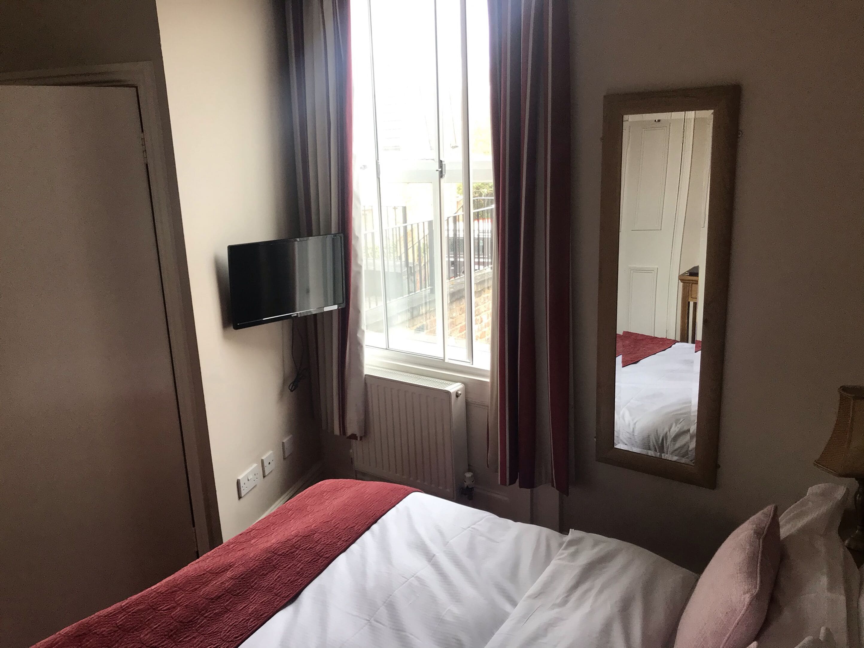 A hotel room with a bed, a television mounted on the wall, a window with curtains, and a mirror reflecting the room.