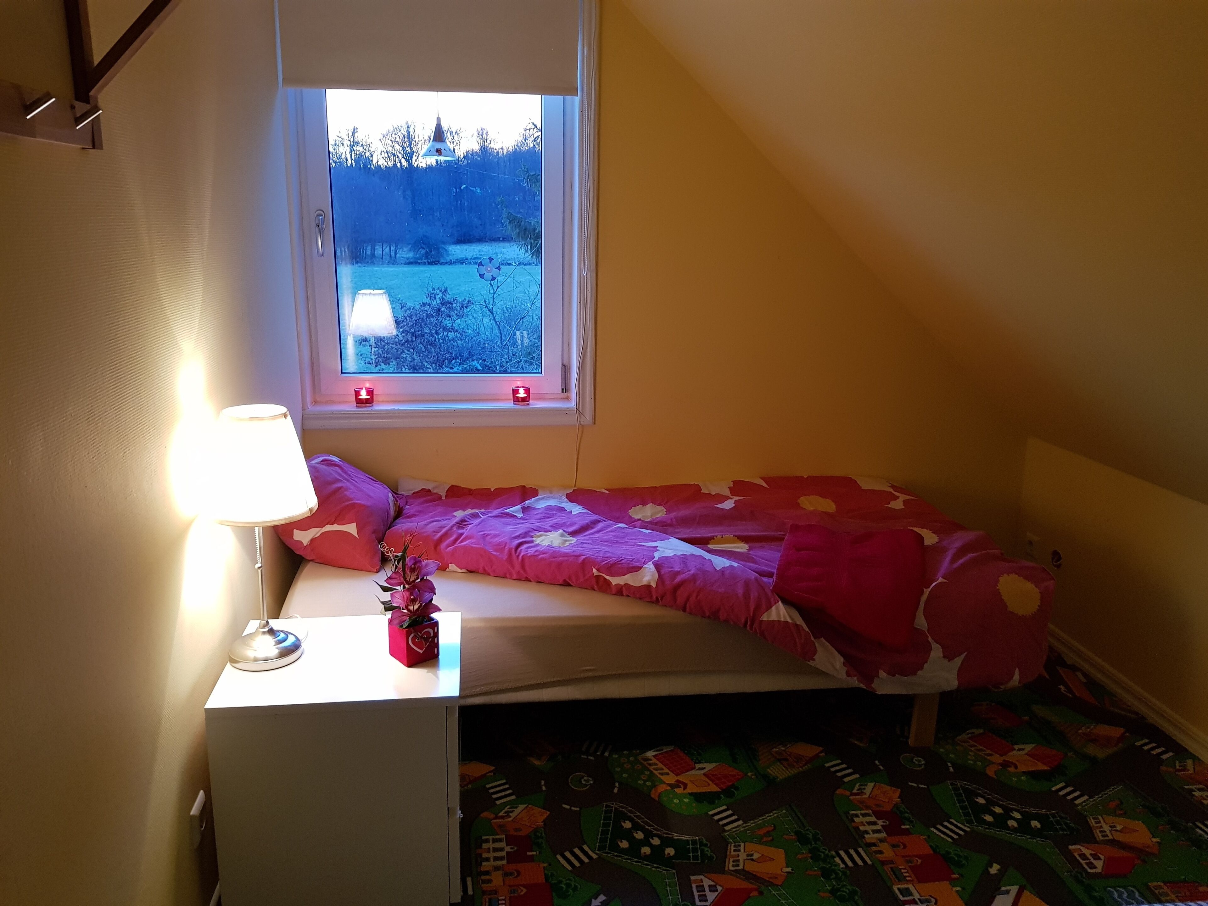 Une petite chambre à coucher avec un lit simple, une table de chevet avec une lampe et une fenêtre donnant sur un paysage enneigé.