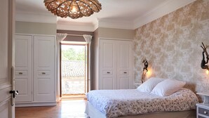 A bedroom with a wooden floor, a bed with a floral bedspread, white curtains, and a decorative wall hanging. - Gîte Le Couvent    . (Cité de Carcassonne)