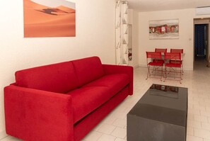 Un canapé rouge, une table basse en verre, un coin repas avec des chaises rouges et un tableau représentant un paysage désertique.
