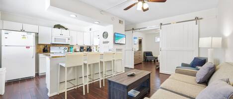 A modern kitchen with white cabinets, a microwave, and a coffee machine. There is a dining area with a wooden table and chairs. The living room has a sofa and a coffee table.