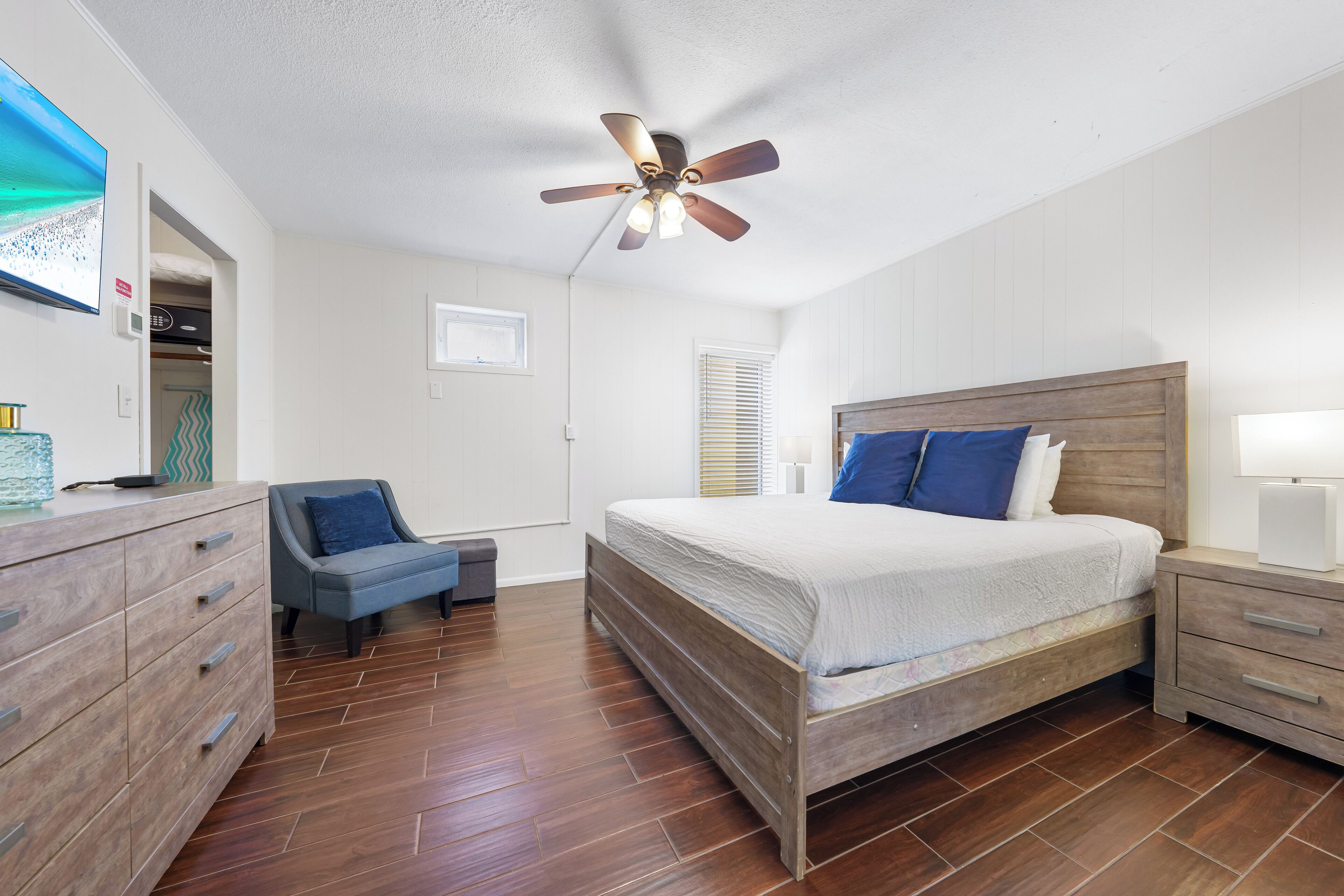 A bedroom with a wooden bed, a nightstand, a dresser, a ceiling fan, and a TV on the wall.