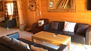 A cozy log cabin interior with a wooden dining table, leather sofas, and a large wall clock.