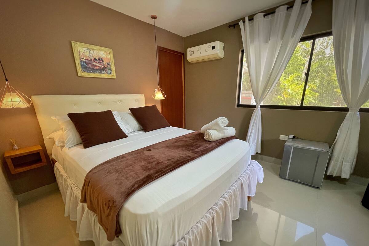 Una cama bien hecha con un plaid marrón, una mesita de noche con lámpara, un cuadro en la pared y una ventana con cortinas.