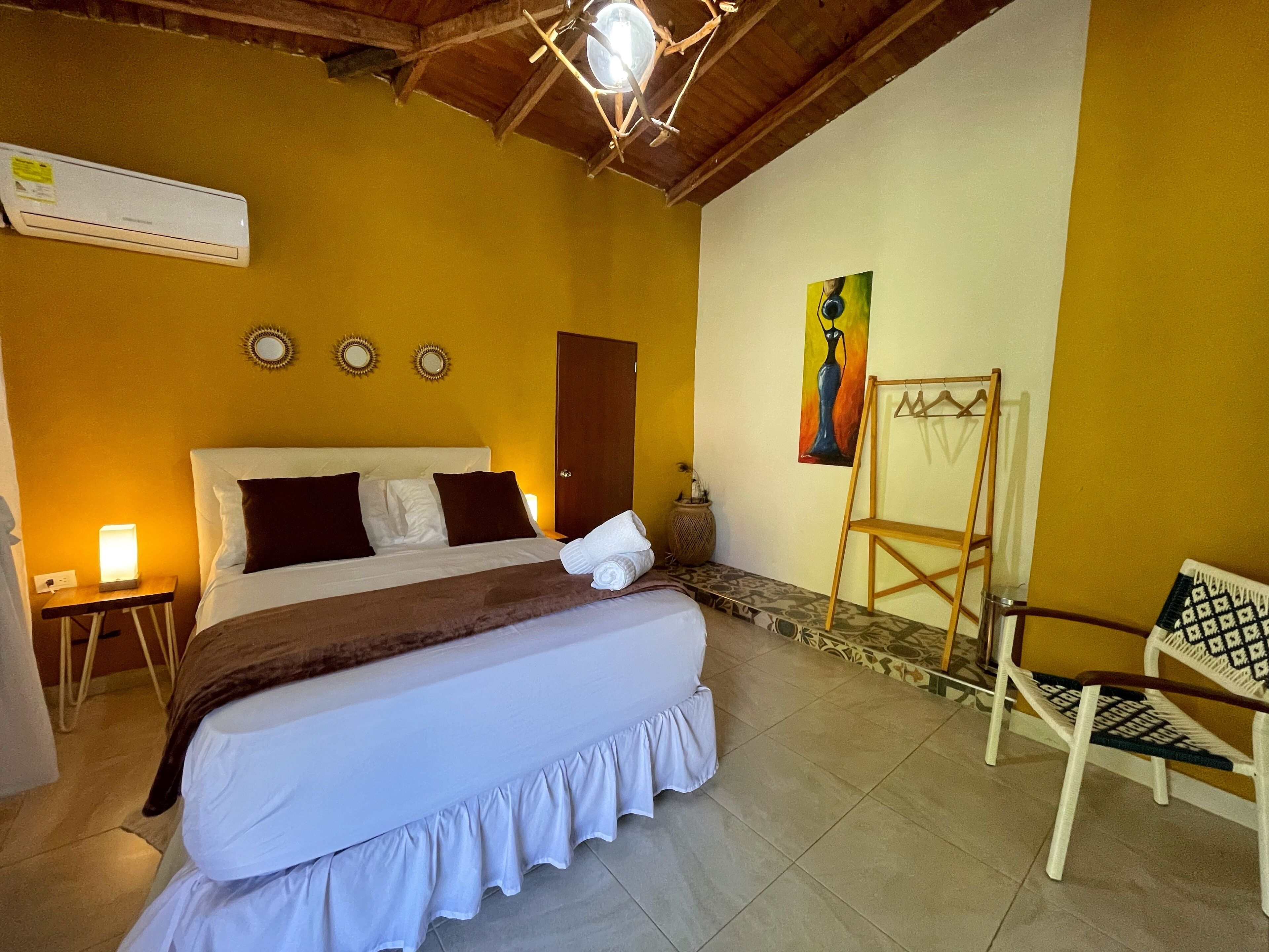 A bedroom with a large bed, a bedside table with a lamp, a wooden ladder, a chair, and a painting on the wall.