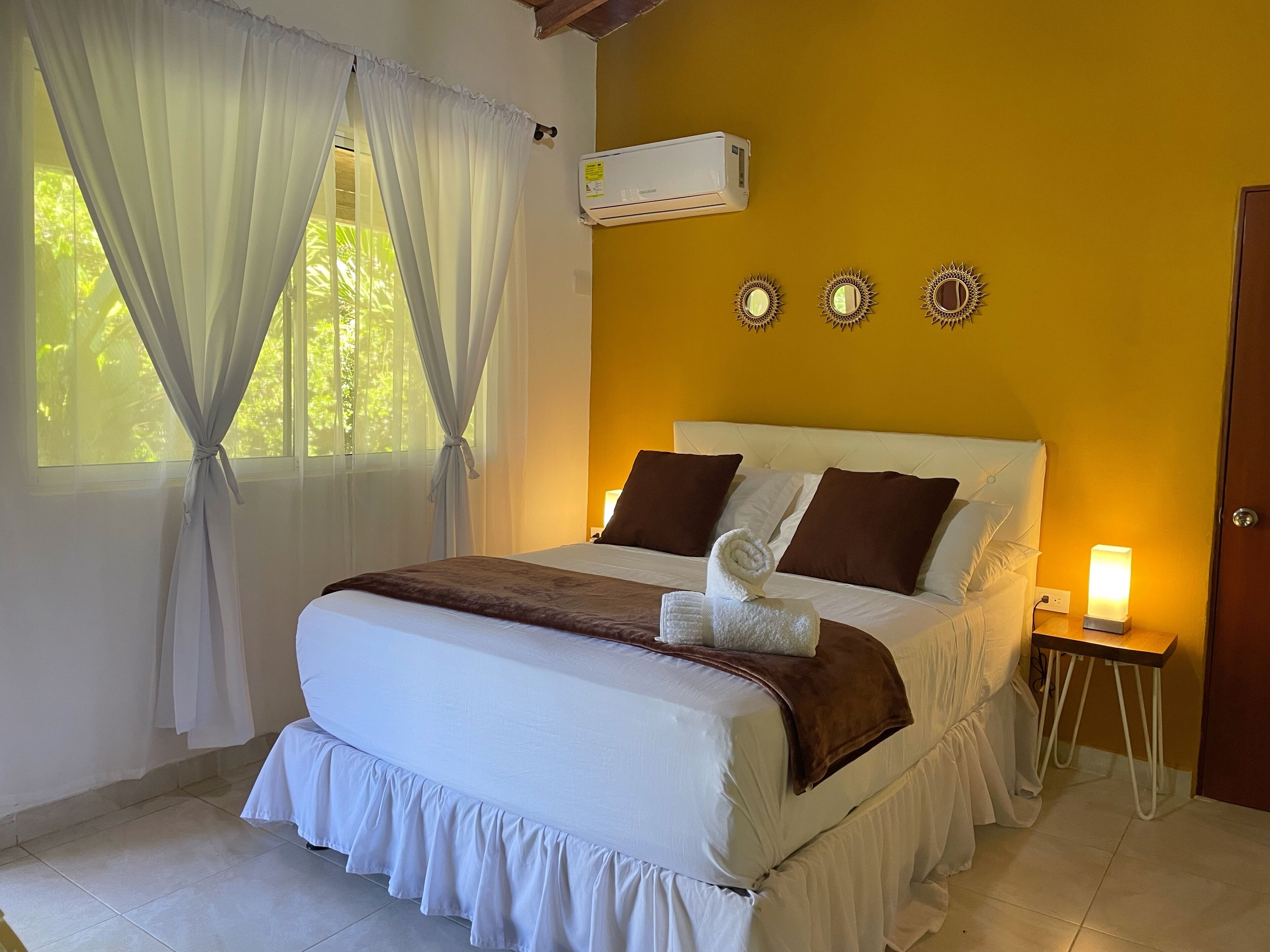 A hotel room with a bed, curtains, an air conditioner, wall decor, and a bedside table with a lamp.