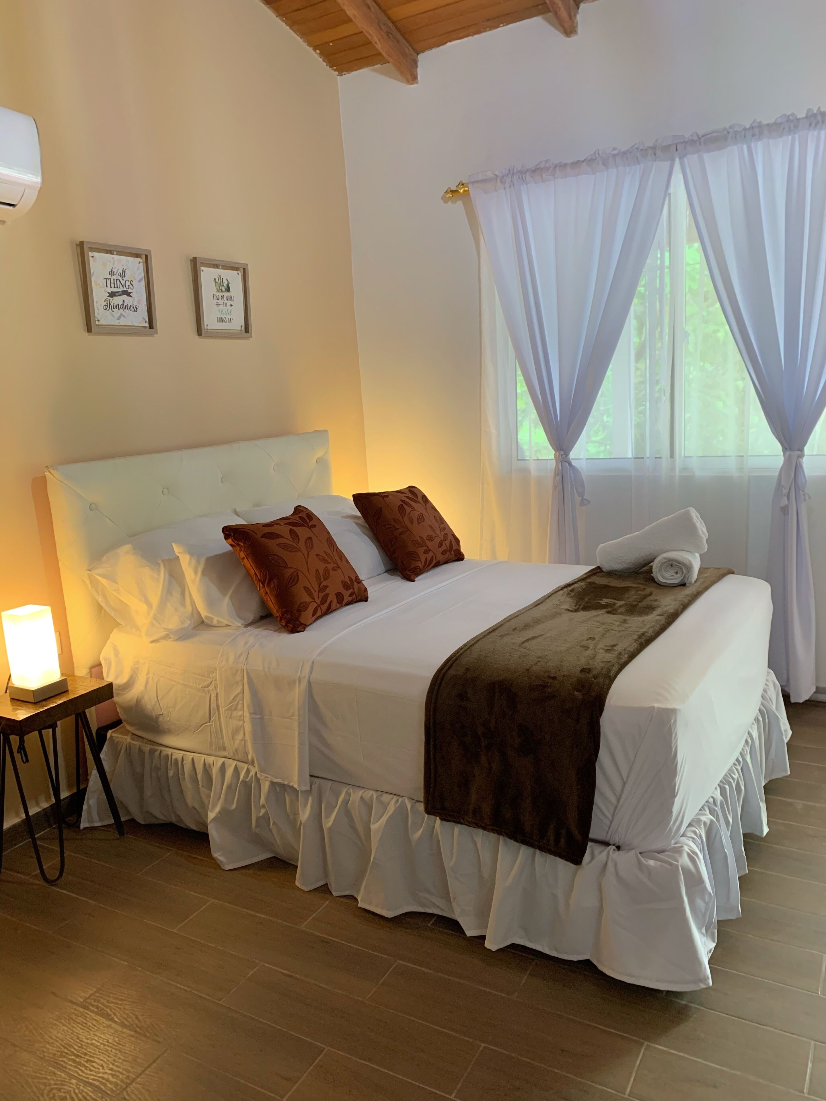 A neatly made bed with a white headboard, two brown pillows, and a brown throw blanket. There is a bedside lamp on a small table beside the bed. The room has wooden flooring and a window with sheer curtains.