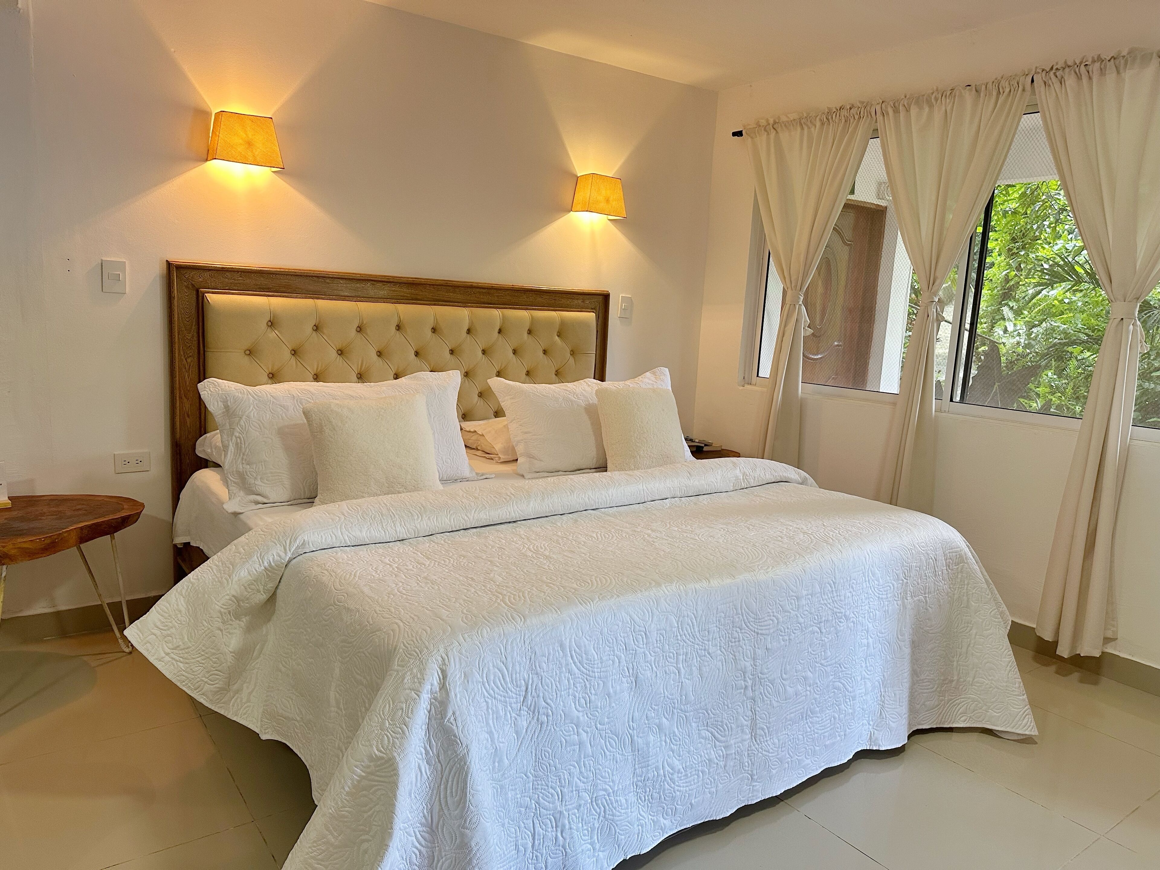 A bedroom with a large bed, two wall-mounted lamps, a wooden headboard, and a window with curtains.