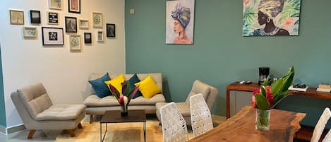 A living room with a sofa, coffee table, dining table, and chairs. There are framed pictures on the wall and a painting above the sofa.