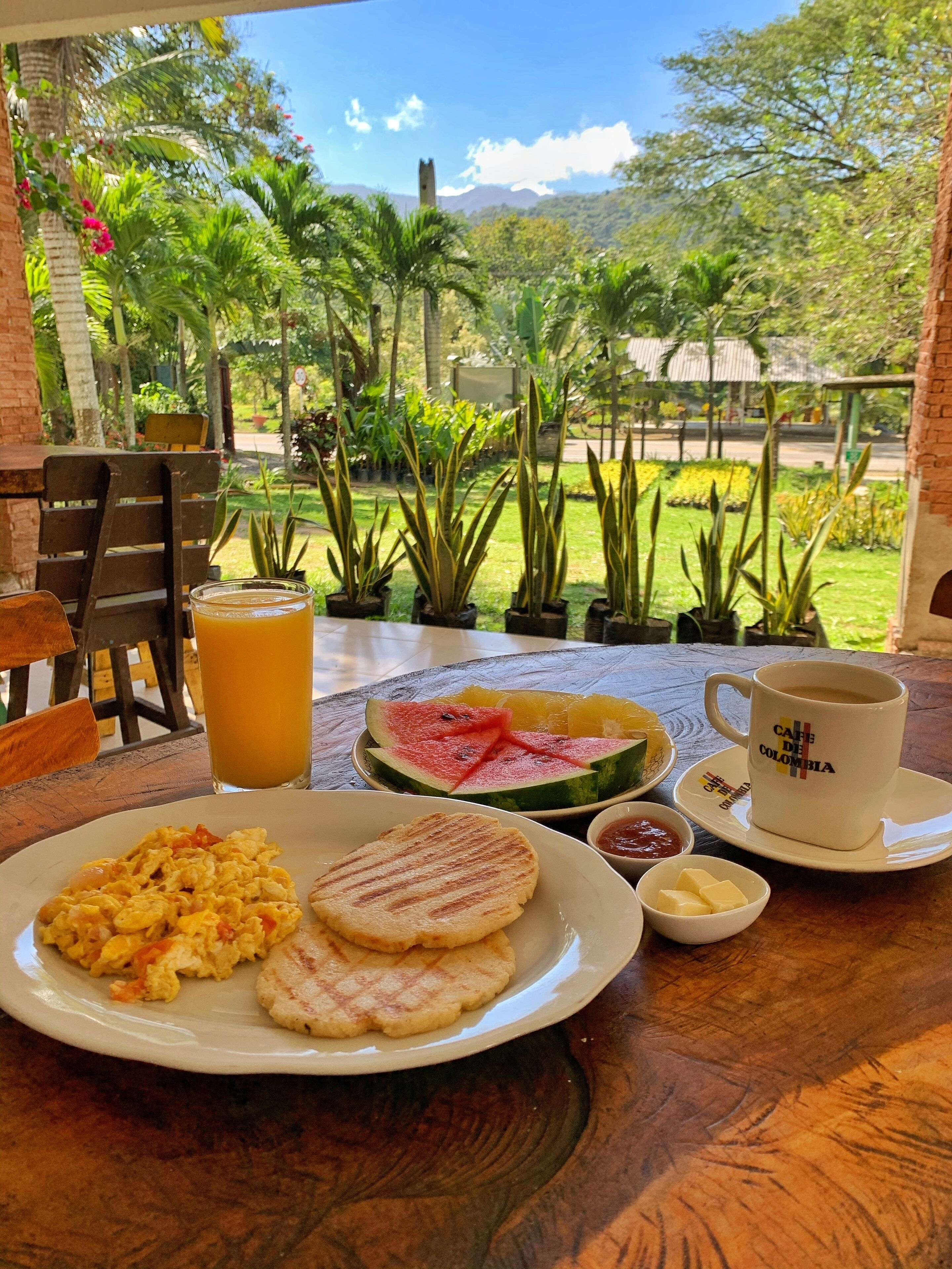 A table set with breakfast including scrambled eggs, grilled bread, a glass of orange juice, a plate of watermelon, and a cup labeled "CAFE DE COLOMBIA".