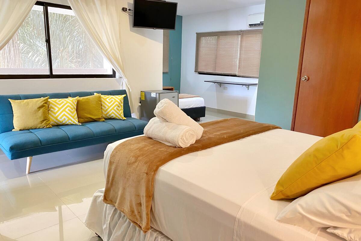 Una habitación de hotel con una cama, un sofá azul, un televisor, una ventana con cortinas y una puerta.
