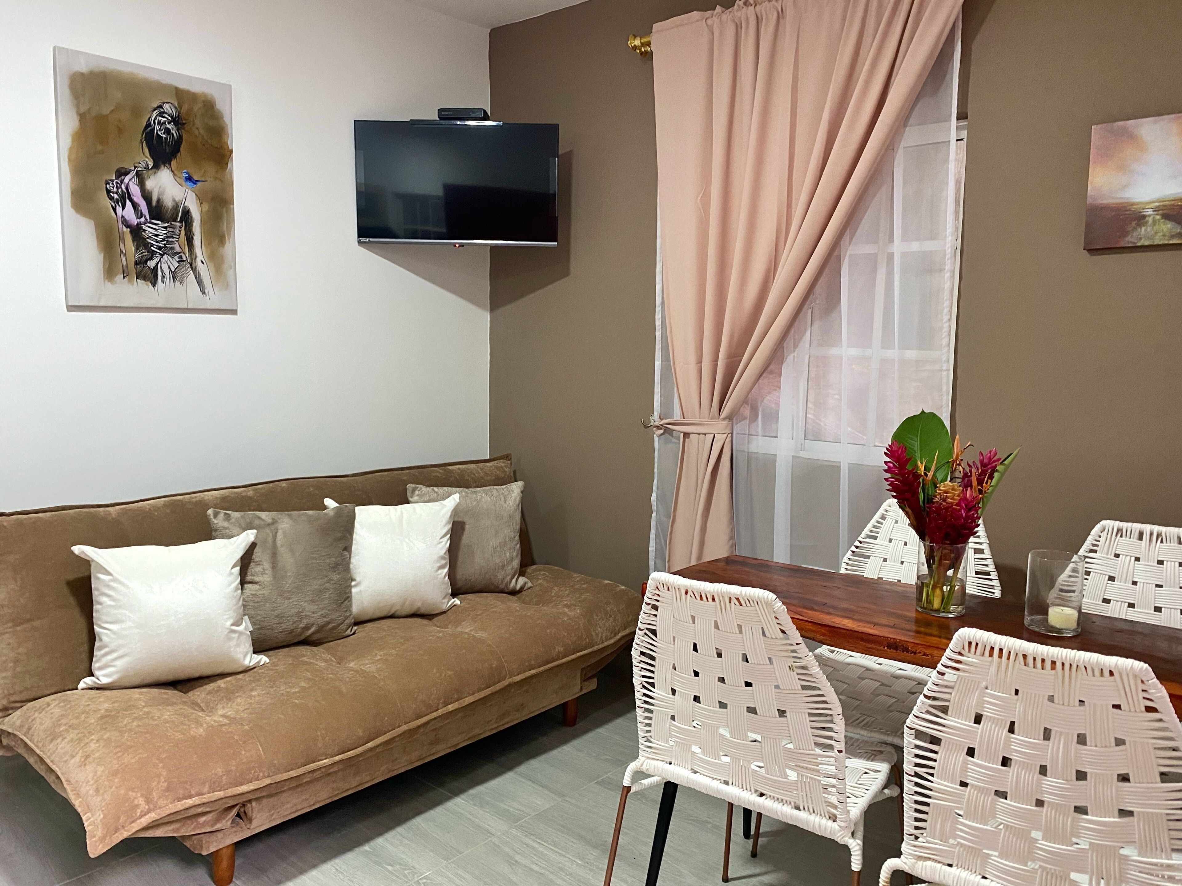 Una sala acogedora con un sofá marrón, cojines blancos, una mesa de comedor de madera y sillas de mimbre blanco.