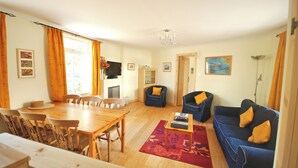 A living room with a wooden dining table, blue sofa, and a TV. - Hydrangea Cottage, Detached 1750,s Stone Cottage, Fully Modernised (Taynuilt)