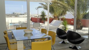 Un espacio exterior moderno para comer con mesa de vidrio y sillas amarillas, con vistas a un paisaje urbano y palmeras.