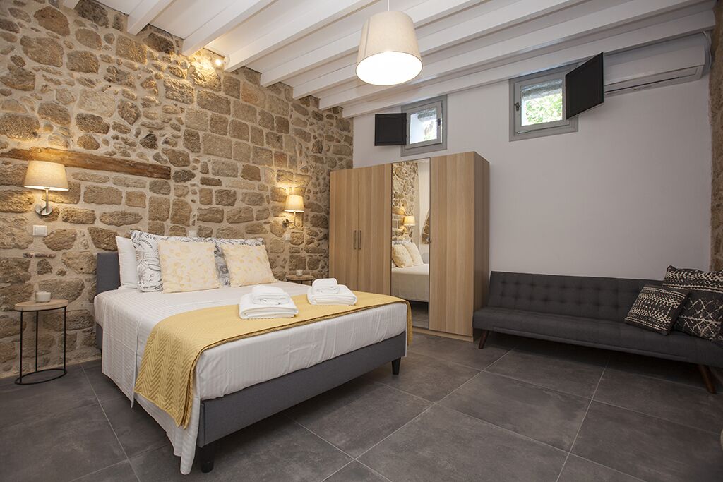 A bedroom with a stone wall, a bed with a yellow bedspread, a grey sofa, and a wooden wardrobe.