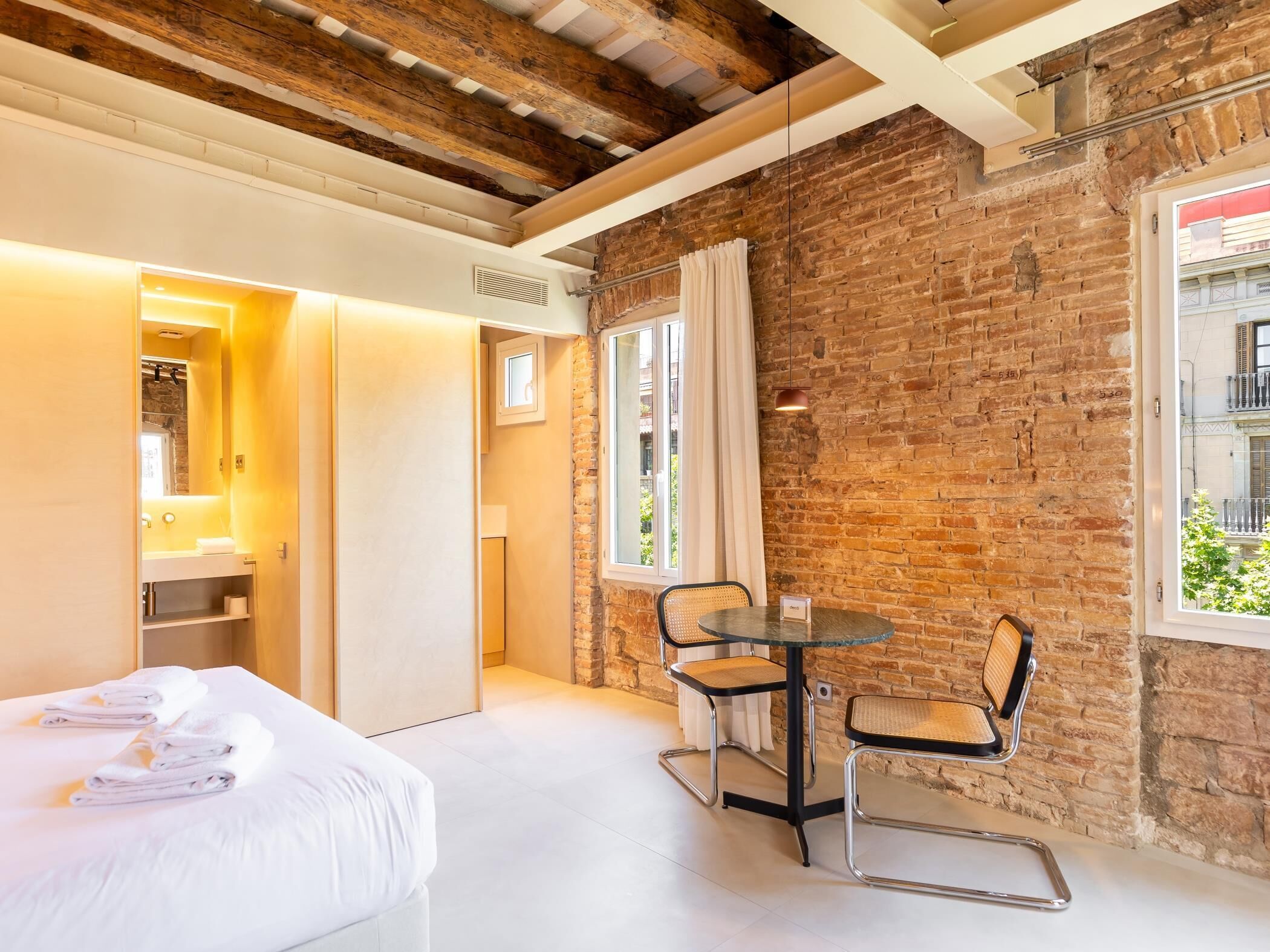Una habitación de hotel moderna con paredes de ladrillo visto, un ventanal con vistas, una cama con toallas ordenadas, una pequeña mesa redonda con sillas y un baño con espejo y lavabo.