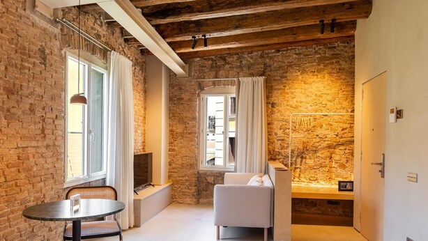 Une chambre d’hôtel avec des murs en briques apparentes, un lit avec du linge de maison blanc, un coin salon avec une table et des chaises, et une fenêtre avec des rideaux.