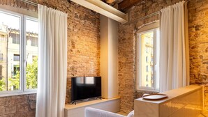 Un salon moderne avec des murs en briques apparentes, un téléviseur à écran plat posé sur une étagère intégrée, un canapé blanc et de grandes fenêtres ornées de rideaux blancs.