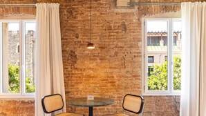 Une pièce avec un mur en briques, deux fenêtres ornées de rideaux blancs, une table ronde accompagnée de deux chaises en osier, et un luminaire suspendu.