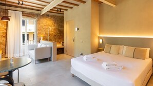 A modern hotel room with a large bed, a sitting area with a table and chair, a stone wall, and a balcony with curtains.