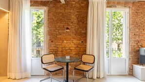 A cozy room with exposed brick walls, a round table with two chairs, and large windows with white curtains.