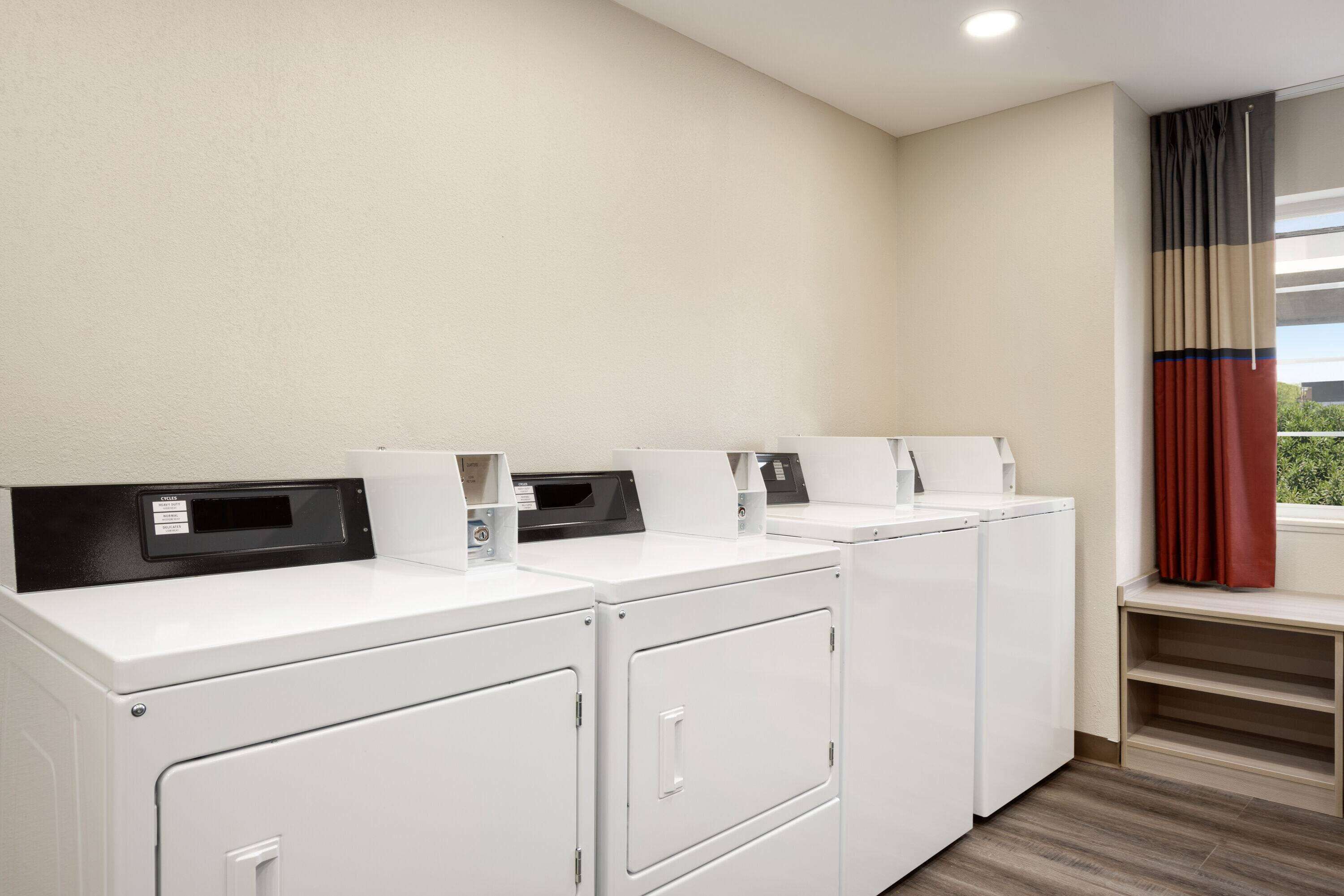 A laundry area with four white washing machines, a control panel, and a window with curtains.