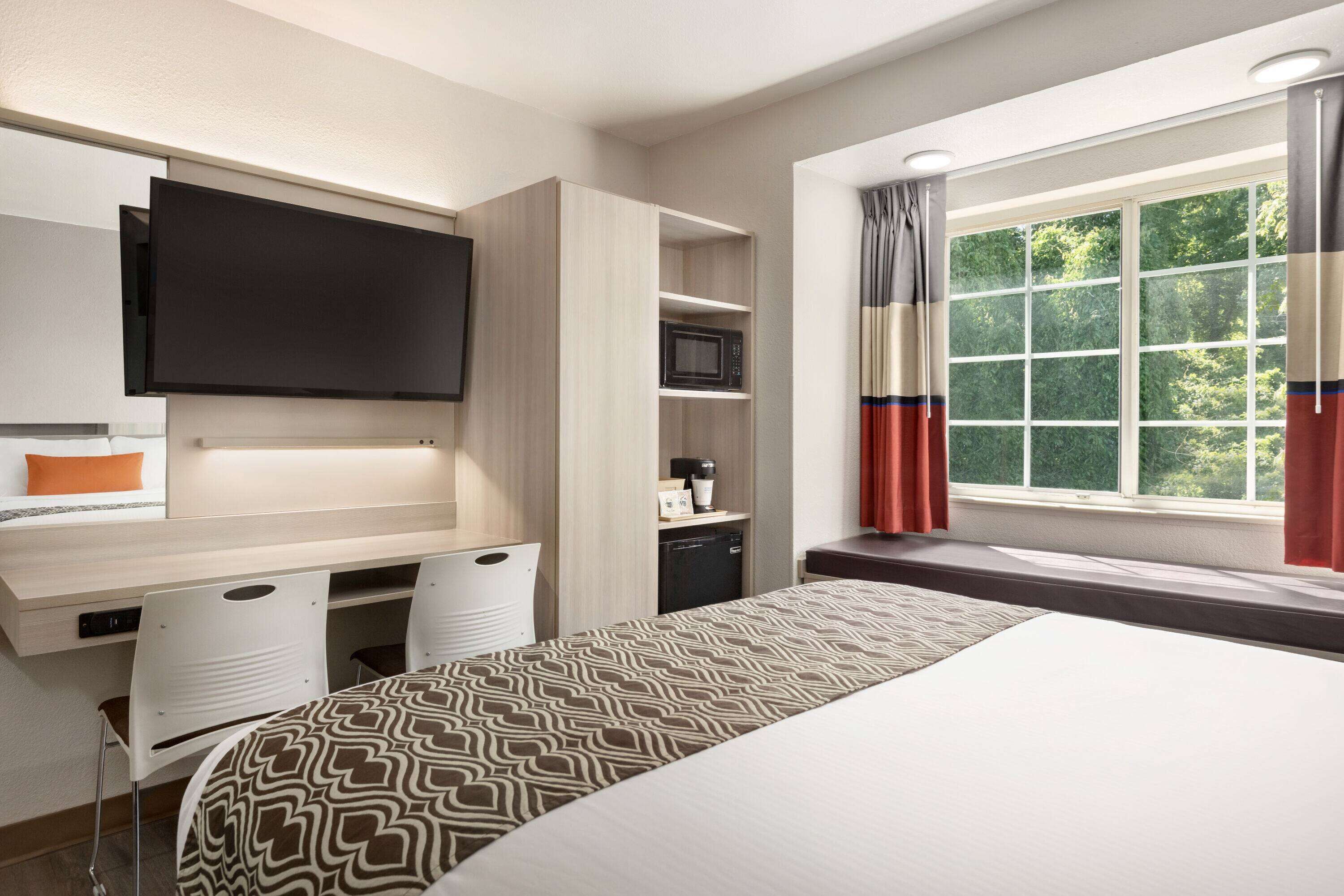 A hotel room with a bed, a desk with a flat-screen TV, a microwave, and a window with curtains.