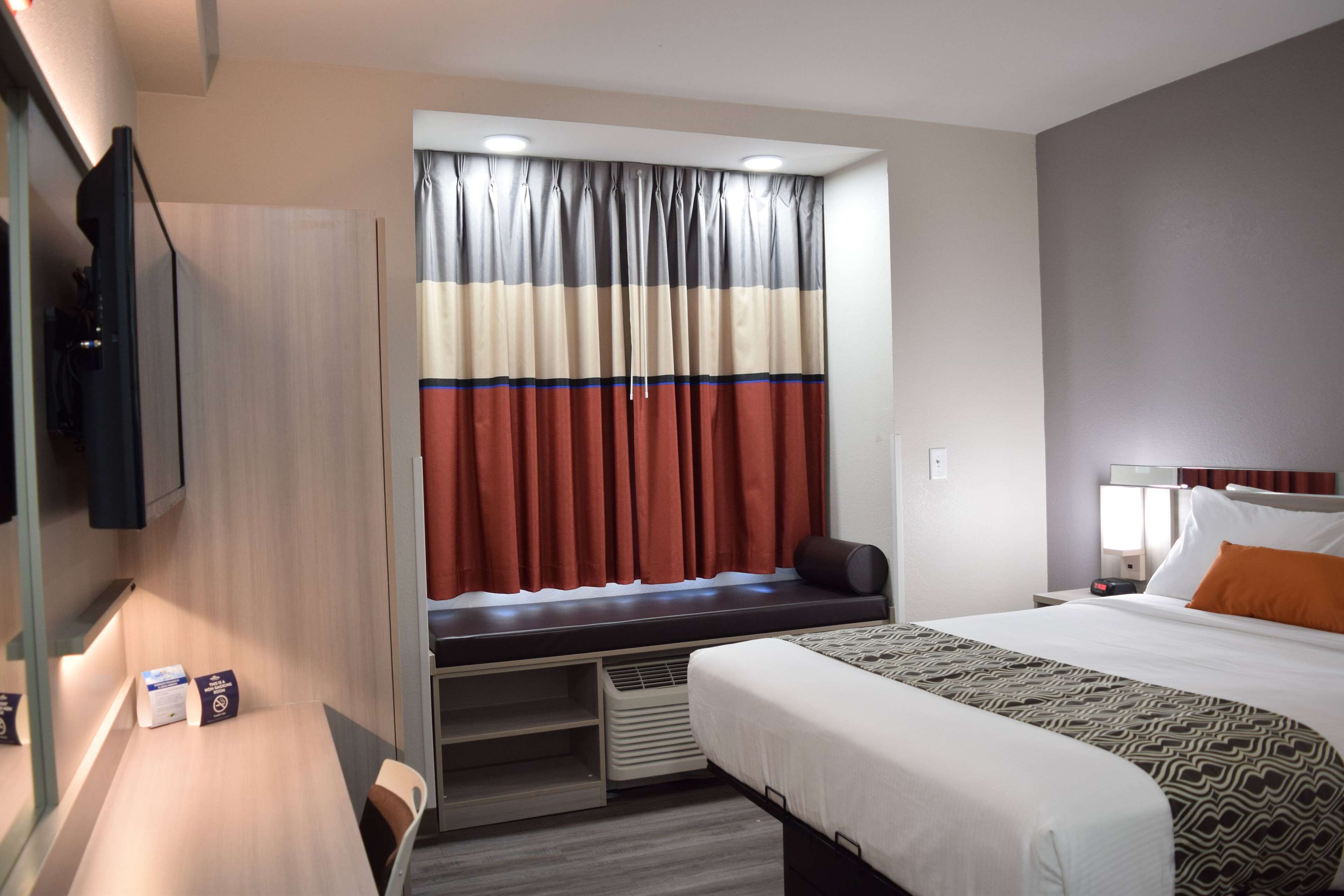 Hotellihuone, jossa on sänky, työpöytä, televisio, ikkuna verhoilla ja sisäänrakennettu penkki.