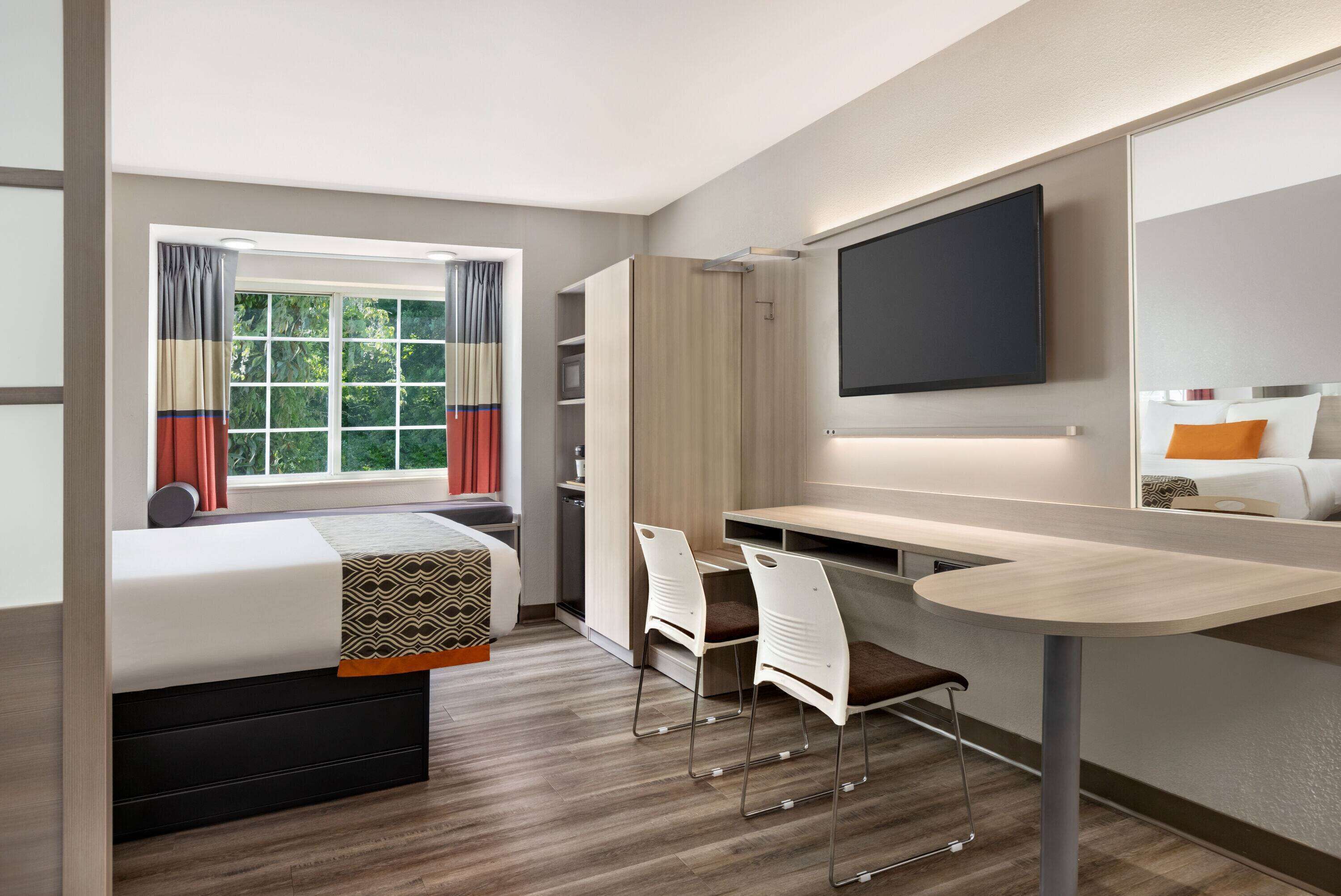 Une chambre d’hôtel moderne équipée d’un lit, d’un bureau, de deux chaises, d’une télévision et d’une fenêtre avec des rideaux.